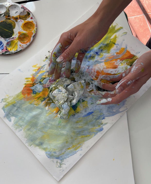 Two paint-covered hands pressing and smearing blue, yellow, orange and white paint directly onto a sheet of paper, with a loaded palette on the table alongside.