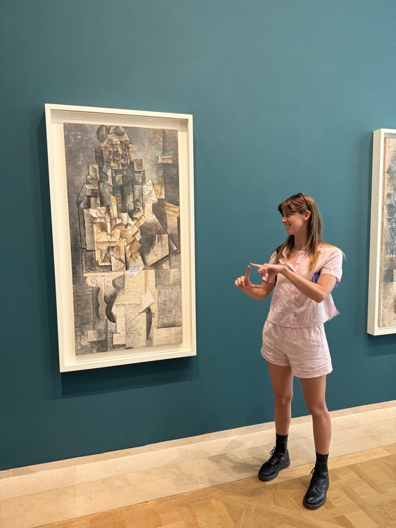 Tamryn standing in a gallery with teal walls, photographing a framed cubist Picasso painting on her phone.