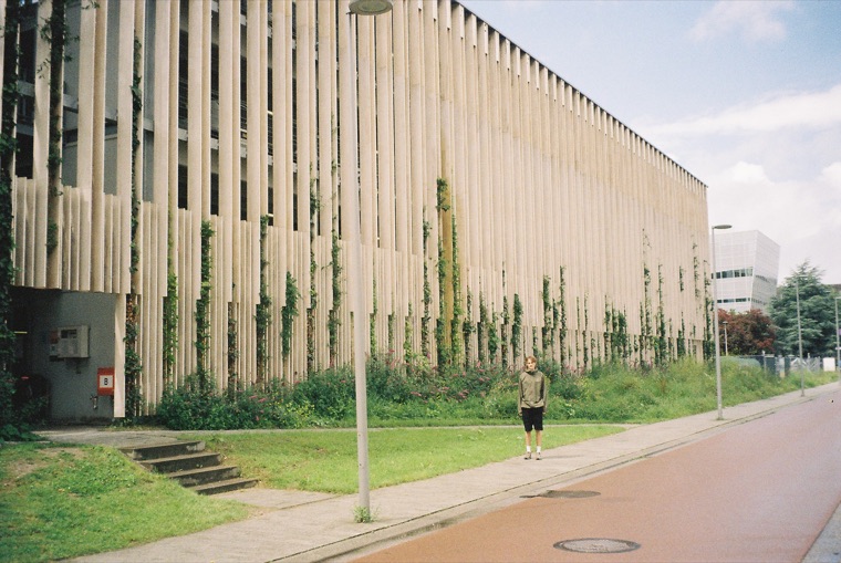 A long concrete building faced with vertical slats, ivy climbing halfway up it, one figure standing on the pavement in front and a street lamp in the foreground.
