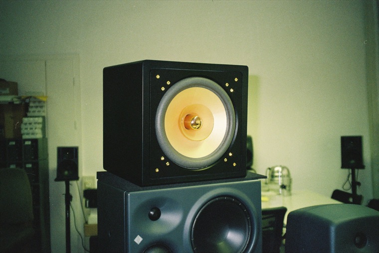 A black loudspeaker cabinet with a large gold-coned driver standing on top of a grey studio monitor, in a room lined with more speakers and equipment racks.