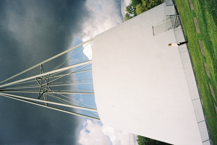 A cable-stayed footbridge photographed with the camera turned on its side, so the pale deck and its fan of white cables run vertically against banked cloud, with one small figure walking on it.