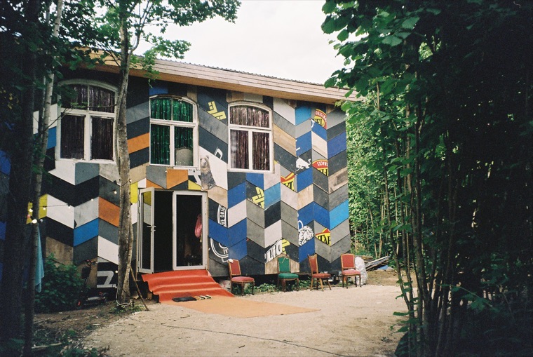 A two-storey house clad entirely in a patchwork of chevrons cut from painted metal and old signage — blues, black, white and orange — with mismatched chairs set out on the sand in front of it.