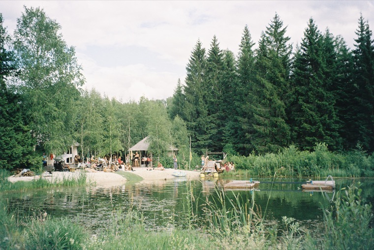 A small forest lake ringed with spruce and birch: people gathered on a sandy beach beside a thatched shelter, and two pedal boats moored in the reeds.