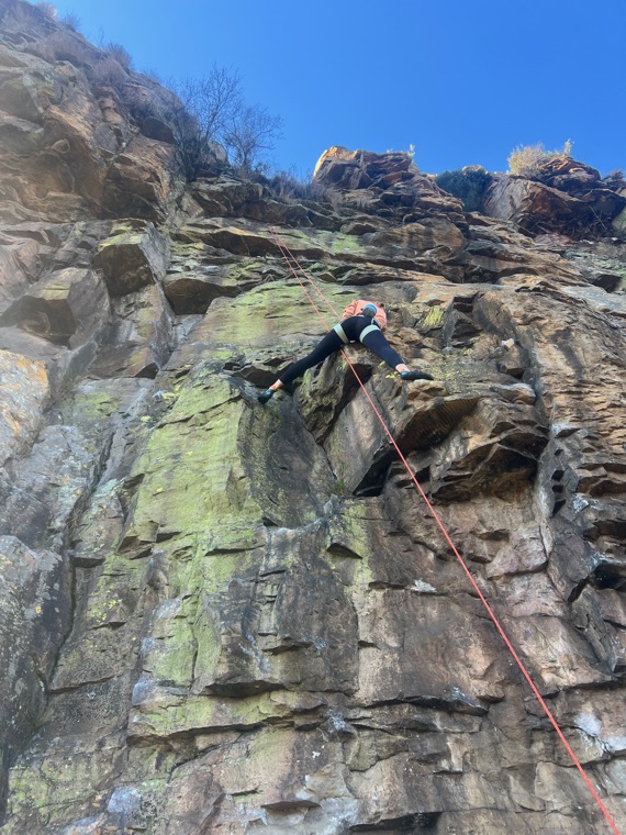 Lead climbing a steep block of lichen-green sandstone under a clear blue sky, the rope running back down the face below.
