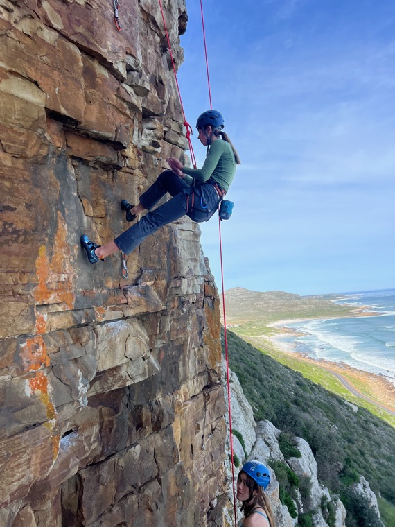 Abseiling down a sandstone face in a helmet and harness, with a long beach and the sea far below.