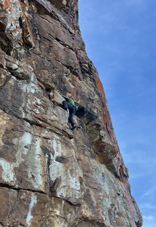 High on a large brown rock face seen from the ground, with two ropes running the whole height of the pitch.