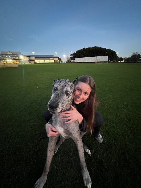 Crouched on a floodlit sports field at dusk with both arms around a large grey merle dog, who is leaning into the camera.