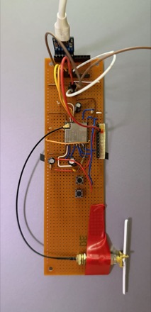 A prototype built on perfboard: a microcontroller module wired in with coloured jumper leads, a radio module at the centre, two push buttons, and a red-bodied antenna connector at the bottom edge.