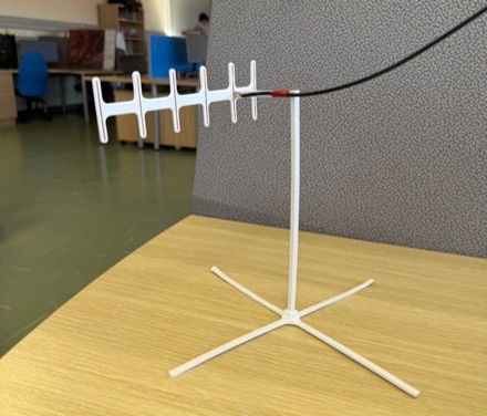 A hand-built white Yagi antenna with six elements, mounted on a printed tripod stand on a desk, with coaxial cable running off to one side.