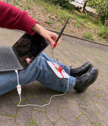 Sitting on a paved path with a laptop balanced on one knee, holding a stub antenna wired to a small red-and-white radio board resting on the other leg.