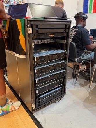 An open server rack holding an Aruba switch above a stack of rack-mounted compute nodes, with team members working at laptops on either side and a flag draped over the table beside it.