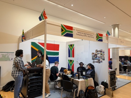 The Centre for High Performance Computing South Africa stand at the conference: a server rack at one end, South African flags pinned across the partitions, and the team working at laptops around a table.