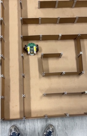 The micromouse part-way through a wooden practice maze photographed from above, with the photographer's shoes at the edge of the board.