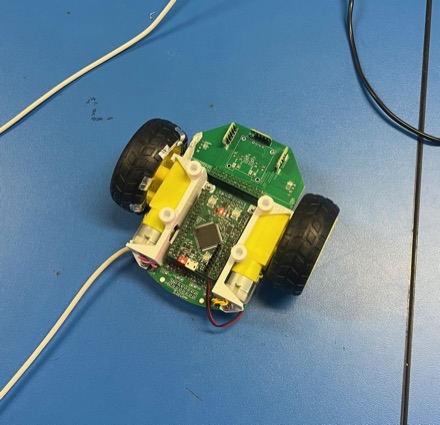 A small two-wheeled robot on a blue mat: a green power supply board stacked over a dark controller board, with two yellow gearmotors and chunky rubber wheels.